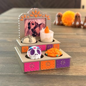 May include: A tiered display stand featuring a photograph of a brown dachshund, a white candle, a small skull with purple and blue accents, and an orange marigold flower. The stand is decorated with colorful panels in pink, orange, and purple.