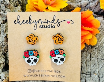May include: A set of four wooden earrings featuring two orange flower designs and two white sugar skull designs with colorful flowers. The earrings are displayed on a brown card with the text "cheeky minds studio" and the social media handle "@cheeky minds" and website "www.cheeky minds.com".