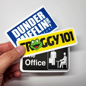 May include: Three stickers featuring the text "Dunder Mifflin, Inc.", "The Office", and "Froggy 101". The "Froggy 101" sticker features a green frog with a yellow background.