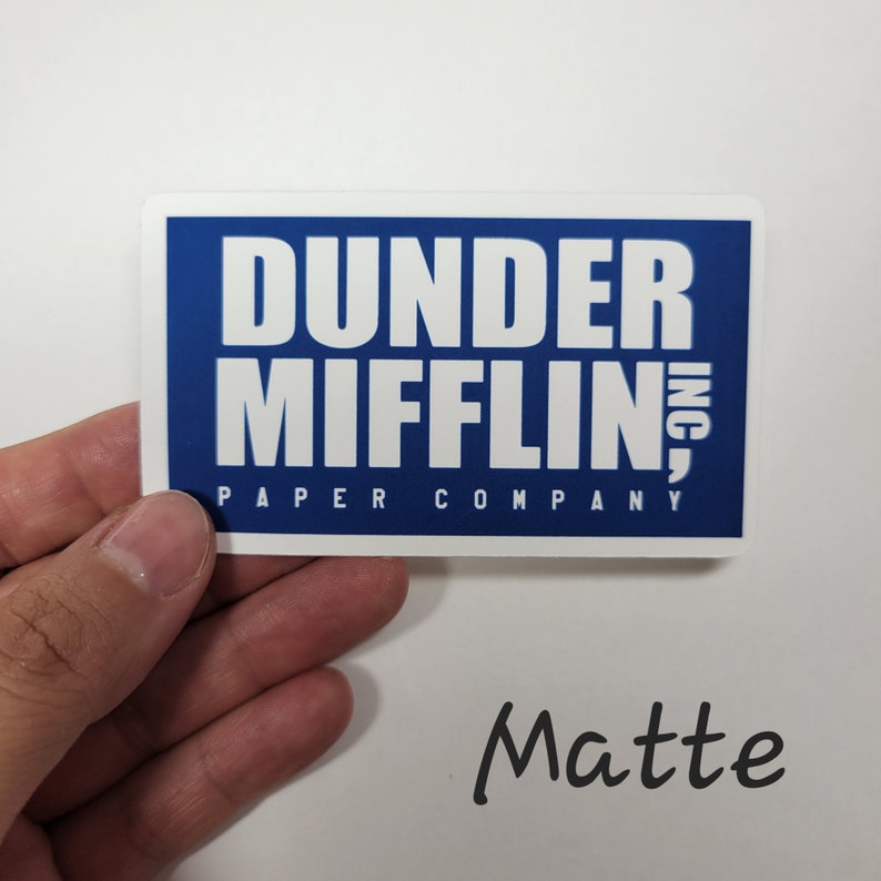 May include: A blue and white sticker with the text "Dunder Mifflin, Inc. Paper Company" in white lettering on a blue background. The sticker has a matte finish.