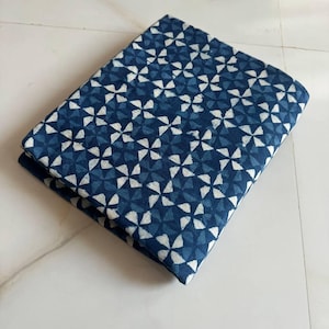 Pode incluir: Um pedaço de tecido azul e branco dobrado com um padrão geométrico. O tecido tem um fundo branco com um padrão repetitivo de cata-ventos azuis.