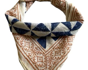 May include: A white bandana with a blue and brown geometric pattern. The bandana is folded and tied in a knot.