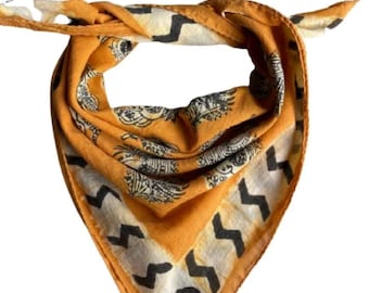 May include: Mustard yellow bandana with a repeating black and white pattern. The bandana features a zig-zag border design and a central motif of stylized floral or animal figures. The fabric is folded and tied.