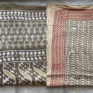 May include: Two folded textiles with printed patterns. One is olive green with white and light green leaf and floral designs. The other is off-white with a red zigzag border and black wavy lines. The fabrics appear to be lightweight.