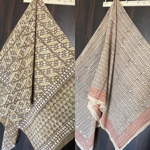May include: Two patterned scarves are displayed. One is brown with a white and olive green leaf design. The other is off-white with a black wavy pattern and a red border. Both are hanging on a white rack.
