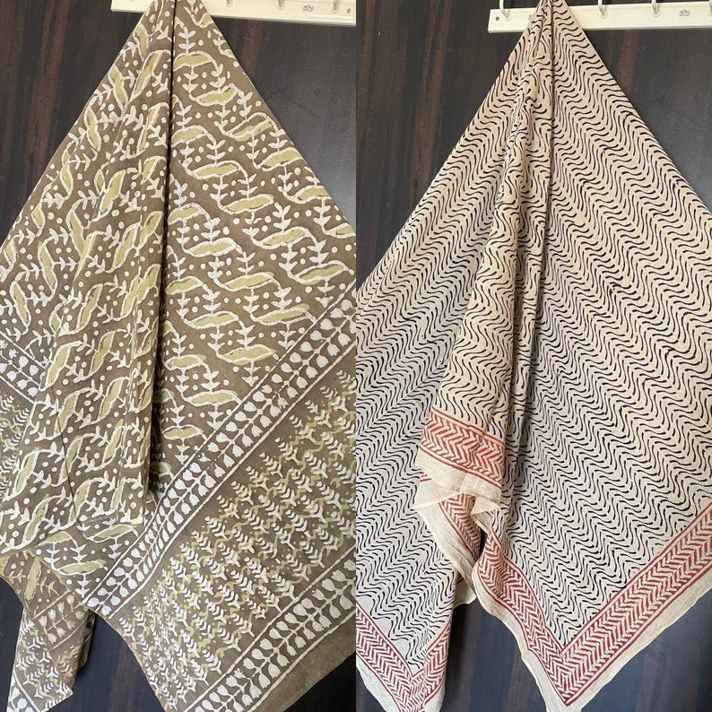 May include: Two patterned scarves are displayed. One is brown with a white and olive green leaf design. The other is off-white with a black wavy pattern and a red border. Both are hanging on a white rack.