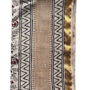 May include: A white cotton fabric with a gold border and a black and brown geometric pattern. The fabric has a floral design on the left side.