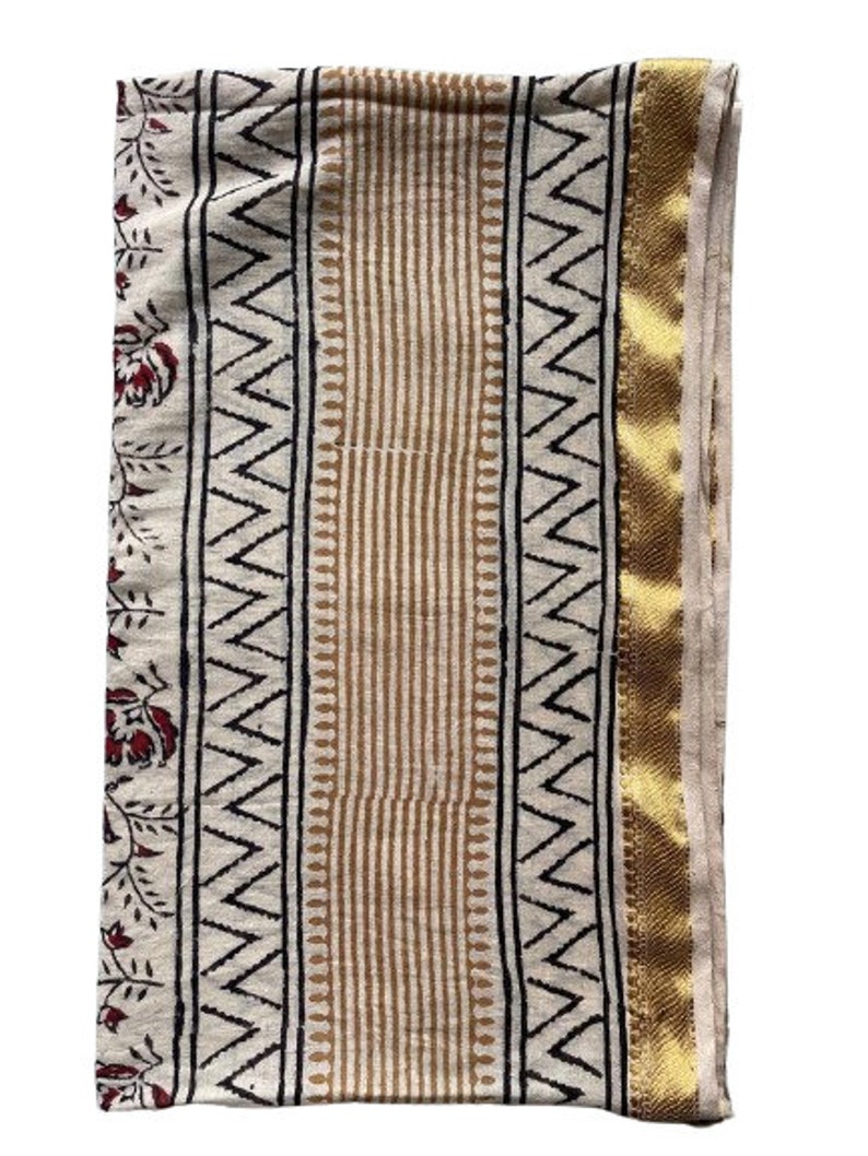 May include: A white cotton fabric with a gold border and a black and brown geometric pattern. The fabric has a floral design on the left side.