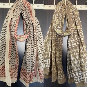 May include: Two patterned scarves hanging on a wall. One is cream-colored with black and red designs, the other is brown with white and light green patterns. Both scarves have a rectangular shape and are made of a lightweight fabric.