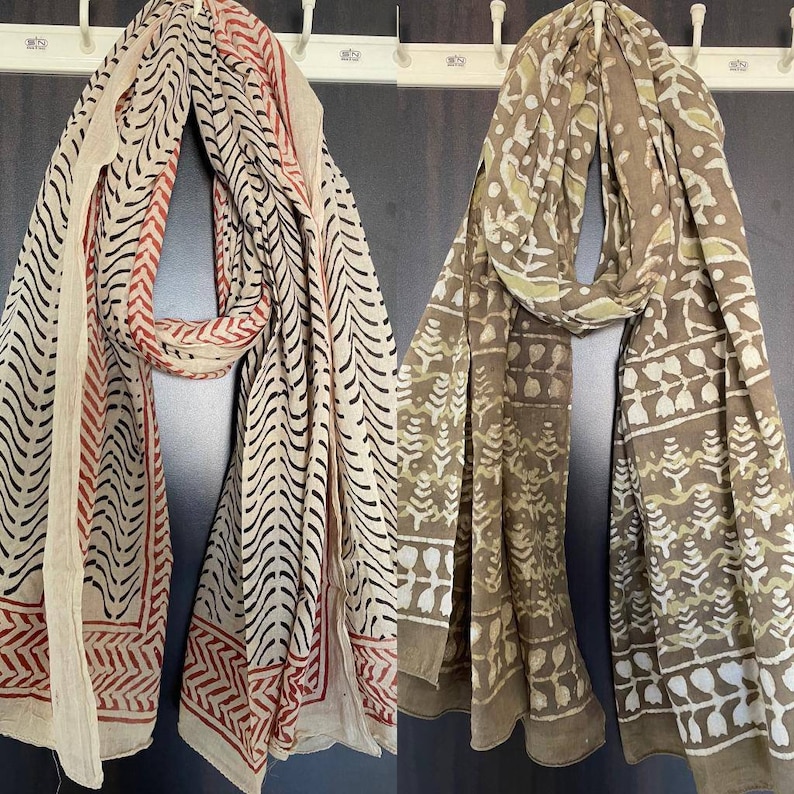 May include: Two patterned scarves hanging on a wall. One is cream-colored with black and red designs, the other is brown with white and light green patterns. Both scarves have a rectangular shape and are made of a lightweight fabric.