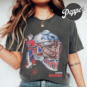 Op de afbeelding: Een antracietgrijs t-shirt met een afbeelding van een ijshockeykeeper die een masker met een berenontwerp draagt. De print bevat een afbeelding van de speler in een rood en blauw uniform, een grote close-up van het masker, een handtekening en de tekst JAKUB DOBES.