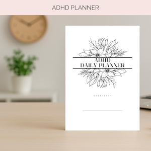 Op de afbeelding: Een witte ADHD Daily Planner met zwarte tekst en bloemenillustraties. De kaft van de planner bevat de tekst "ADHD Daily Planner" en de jaren 2025/2026. De planner ligt op een lichtgekleurd houten oppervlak.