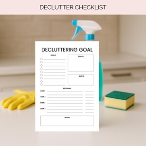 Op de afbeelding: Een witte checklist voor het opruimen met de titel "DECLUTTERING GOAL" en de tekst "DECLUTTER CHECKLIST" bovenaan. De checklist bevat secties voor doelen, focus, een citaat en notities. Een gele schoonmaakhandschoen, een groene en gele spons en een blauwe spuitfles staan op de achtergrond.