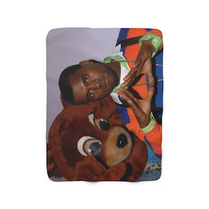 May include: A man wearing a blue and orange shirt with a green and gold chain is posing with a large brown bear plush toy. The man is making a heart shape with his hands.
