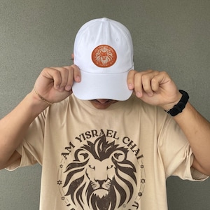Puede incluir: Una gorra de béisbol blanca con un logotipo de león naranja en la parte delantera. La gorra la lleva una persona que lleva una camiseta beige con un gráfico de león marrón y el texto "Am Yisrael Chai" en hebreo.