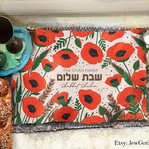 May include: A floral placemat with red poppies and green leaves, with the text "The Stern Family Shabbat Shalom." A braided loaf of bread, a wine bottle, and a blue ceramic wine holder are also visible.