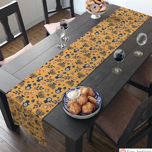 Puede incluir: Un camino de mesa amarillo con un patrón azul y blanco de manos de hamsa, menorás y granadas. El camino de mesa está sobre una mesa de madera oscura con un tazón de donas y un plato de comida frita.