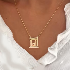 Può includere: Una collana d'oro con un ciondolo a forma di edificio con un cuore al suo interno. La collana è indossata da una persona con una camicia bianca.