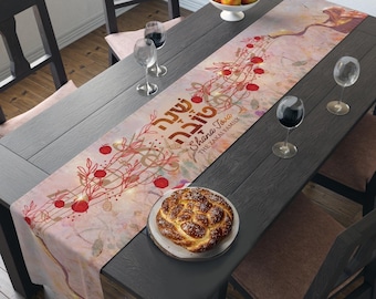 May include: A pink and gold table runner with a floral design and red apples. The text "Shana Tova" and "The Zakai Family" is printed on the runner.