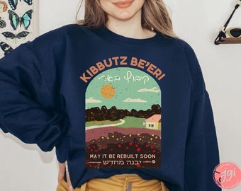 Op de afbeelding: Een donkerblauw sweatshirt met een grafisch ontwerp met een landschapsscène met een huis, bomen en een zon. De tekst "Kibbutz Be'eri" staat in een cirkelvormig ontwerp boven de scène, met de Hebreeuwse tekst "קיבוץ בארי" in de cirkel. De tekst "May it be rebuilt soon" staat onder de scène, met de Hebreeuwse tekst "בְּעִבְרִית מְהַר" eronder.