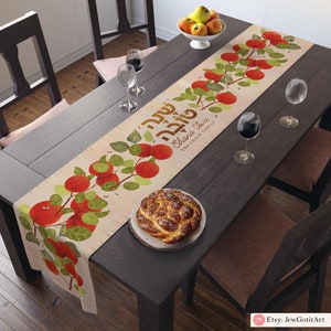 May include: A beige table runner with a red apple and green leaf design. The runner features the Hebrew text "Shana Tova" and the words "The Chain Family".
