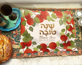 Könnte beinhalten: Ein weißer Tischläufer mit einem roten Apfelmuster und den hebräischen Worten "Shana Tova" in goldener Schrift. Der Text "The Chaim Family" ist ebenfalls in goldener Schrift unter dem hebräischen Text geschrieben.