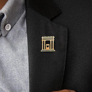 Può includere: Una spilla da bavero dorata che presenta una rappresentazione stilizzata di un edificio con un cuore al suo interno. La spilla è indossata su un blazer nero.