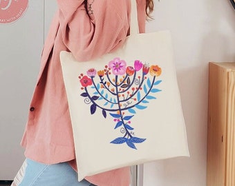 Könnte beinhalten: Eine weiße Canvas-Tragetasche mit einem farbenfrohen Blumendesign, das eine Menora zeigt. Die Tasche hat einen rosafarbenen Trageriemen und der Text "Jew Got It" ist auf die Tasche gedruckt.