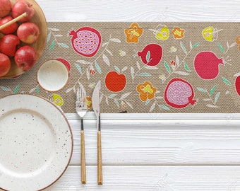 Könnte beinhalten: Ein weißer Tisch mit einem braunen und beigefarbenen Tischläufer mit einem Granatapfel-Design. Der Läufer hat rote, orange, gelbe und rosa Granatäpfel mit grünen Blättern. Auf dem Tisch liegen ein weißer Teller, eine Gabel und ein Messer.
