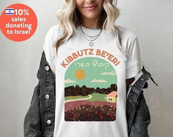 May include: White t-shirt with a graphic design of a landscape with a house and the text "Kibbutz Be'eri" in Hebrew and English.