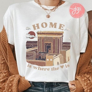 May include: White t-shirt with a graphic illustration of a building and the text "HOME is where the heart is". The graphic is in shades of brown, tan, and burgundy. The shirt is worn with blue jeans and a brown cardigan.