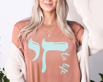 Op de afbeelding: Een lichtbruin T-shirt met een blauwe Hebreeuwse letter en de woorden "to life" in blauw cursief.