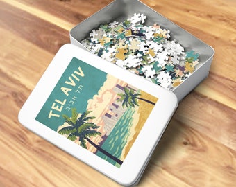 Op de afbeelding: Een witte metalen doos met een kleurrijke illustratie van Tel Aviv, Israël, op de deksel. De doos is open en gevuld met witte puzzelstukjes. De tekst op de deksel luidt "Tel Aviv" in het Engels en Hebreeuws.