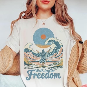 Puede incluir: Camiseta blanca con un gráfico de una persona caminando hacia una ola con el texto "Walk into Freedom".
