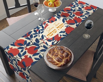 May include: A table runner with a pomegranate design and the text "Shana Tova" in Hebrew and English. The runner is on a dark wood table with a challah bread and two glasses of wine.