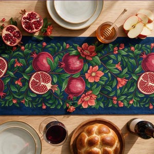 May include: A rectangular table runner featuring a dark blue background with a repeating botanical pattern of red pomegranates, green leaves, and pink flowers. The design is printed across the fabric, showcasing whole and halved pomegranates.