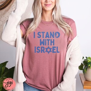 May include: A mauve t-shirt with the text "I Stand With Israel" in blue lettering. The text includes a blue silhouette of a person and a blue Star of David.