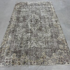 Puede incluir: Una alfombra vintage con un aspecto descolorido y desgastado. La alfombra tiene un fondo beige claro con un patrón marrón oscuro y gris. La alfombra está hecha de lana y tiene una textura anudada a mano.