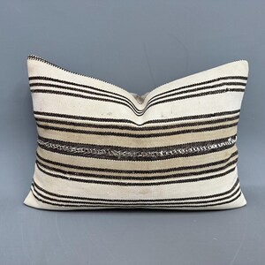 May include: Cream-coloured rectangular cushion featuring horizontal stripes in black and tan. The cushion has a woven texture and a slightly worn appearance, suggesting a vintage or handmade origin. The stripes vary in width.