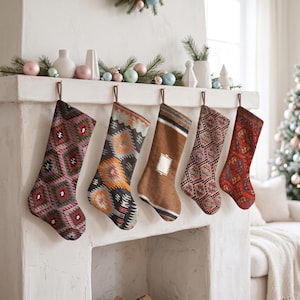 Può includere: Cinque calze natalizie appese in fila su un camino bianco. Le calze presentano vari motivi intrecciati, tra cui forme geometriche, rombi e strisce nei colori rosso, verde, grigio, arancione, marrone e crema con passanti marroni.