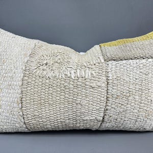 May include: A white and beige woven pillow with a yellow and white stripe on one side. The pillow is made of a textured fabric and has a slightly distressed look.