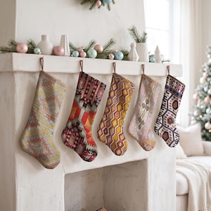 Puede incluir: Cinco calcetines de Navidad colgados en fila de una repisa de chimenea blanca. Cada calcetín presenta un patrón tejido distinto con paletas de colores variadas que incluyen amarillo, rojo, naranja, morado, azul y crema, con lazos marrones.