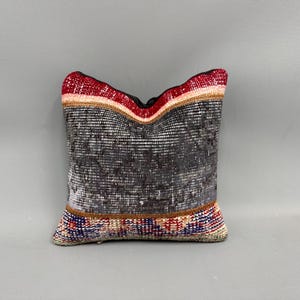 May include: Square decorative pillow with a distressed, textured design. The pillow features a mix of gray, black, red, and beige tones. The top edge has a red border, and the bottom edge has a colorful striped pattern. The pillow is likely made of woven fabric.