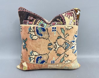 May include: A square decorative pillow with a vintage rug design. The pillow features a peach-colored background with floral patterns in blue, green, and white. The top and bottom edges have a darker, patterned border in shades of brown, pink, and beige.