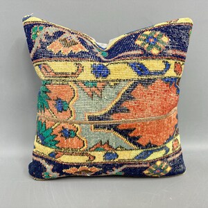 May include: Square decorative pillow with a vintage rug design. The pillow features a patchwork pattern with a navy blue border, yellow, orange, and teal floral motifs. The pillow is a square shape.