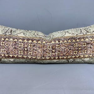Puede incluir: Cojín rectangular decorativo con diseño de alfombra vintage. El cojín presenta una banda central de patrones geométricos marrones y beige, enmarcada por un patrón floral verde claro y beige. De forma alargada.