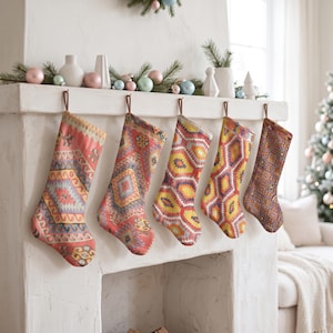 Könnte beinhalten: Fünf Weihnachtsstrümpfe hängen in einer Reihe an einem weißen Kaminsims. Jeder Strumpf weist ein ausgeprägtes geometrisches Webmuster in leuchtenden Farben wie Rot, Orange, Gelb, Blau und Violett auf und ist an braunen Schlaufen aufgehängt.