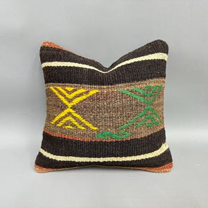 May include: Square decorative pillow with a woven design. The pillow features a dark brown background with horizontal stripes and geometric patterns in yellow and green. The pillow is approximately 16 inches square.