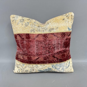 May include: Square decorative pillow with a patchwork design. The pillow features a central band of deep red, bordered by sections of beige and cream with faded floral patterns. The overall aesthetic is vintage and textured.