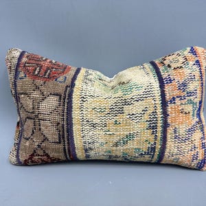 May include: A rectangular pillow with a colorful patterned fabric. The fabric is a mix of beige, brown, blue, and orange. The pillow is made from a woven fabric with a geometric design.